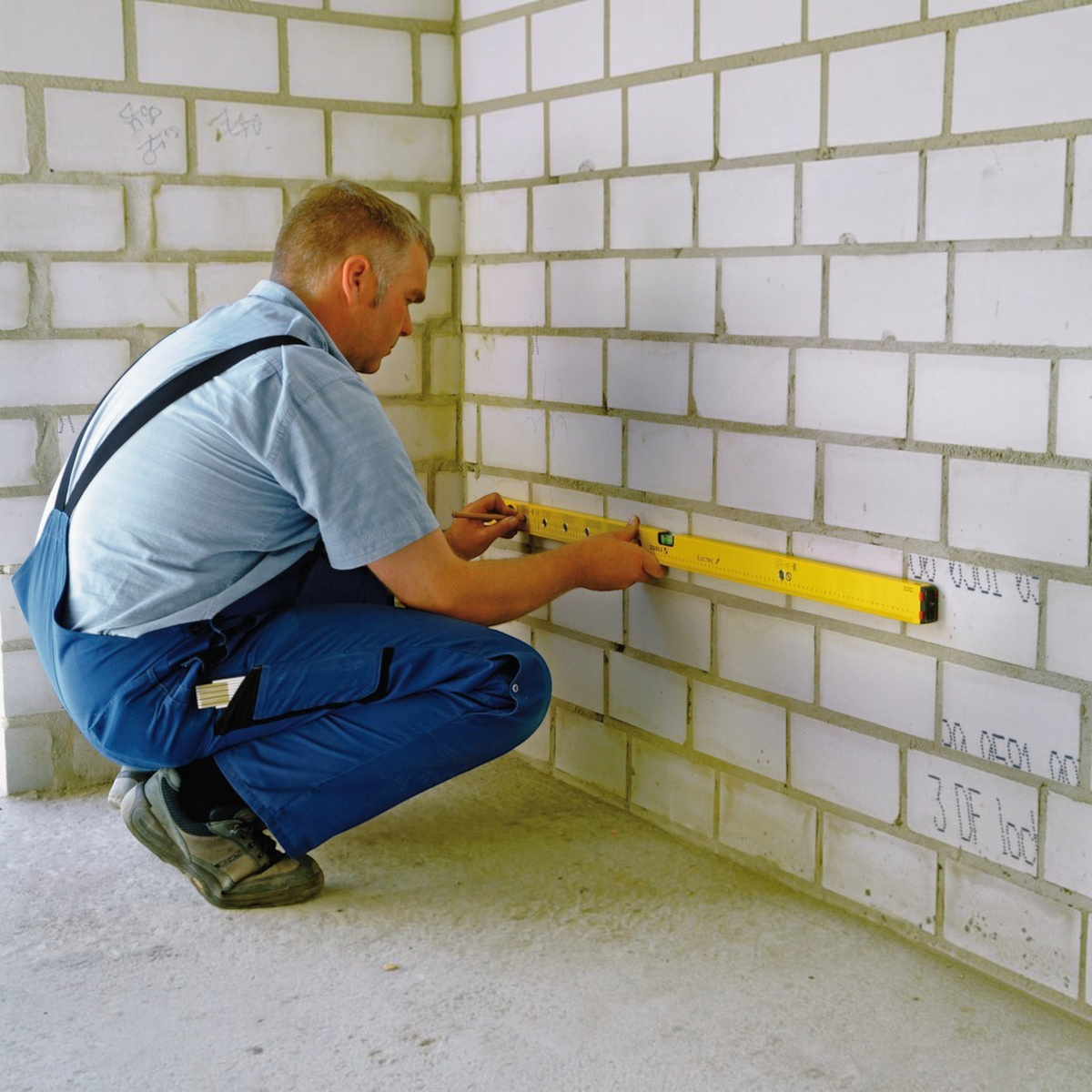 Handwerker von Werkfit nutzt eine gelbe Wasserwaage Type 70 Electric zum Anzeichnen an einer weißen Ziegelwand