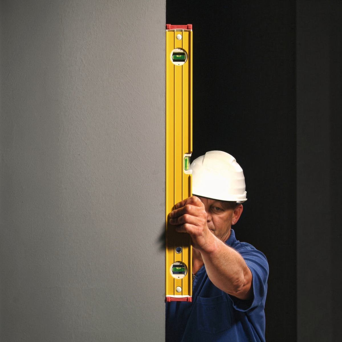 Handwerker von Werkfit benutzt eine gelbe Wasserwaage Type 96-2 an einer grauen Wand mit Schutzhelm